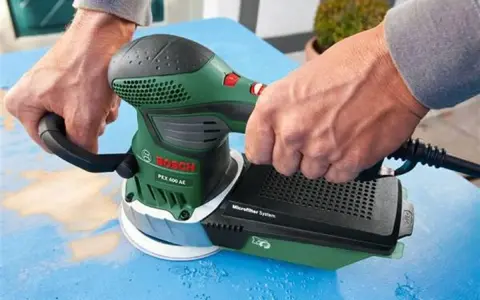 Dłonie trzymają zieloną szlifierkę Bosch PEX 400 AE. To szlifierka oscylacyjna a mimośrodowa, która usuwa starą farbę z niebieskiej powierzchni.