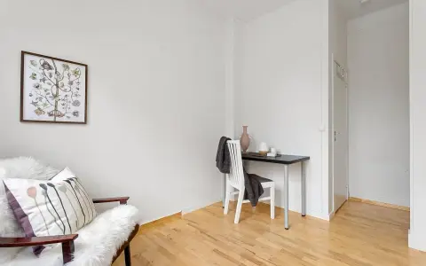 Jasna przestrzeń z fotelem, obrazem botanicznym i małym biurkiem. Wazon i świeca na blacie, a na krześle narzucony koc. Kolory nawiązują do palety Pantone.