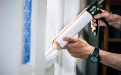 Ręce trzymają pistolet do silikonu, aplikując go na ramę okna. To pokazuje, co to jest silikon – materiał uszczelniający.