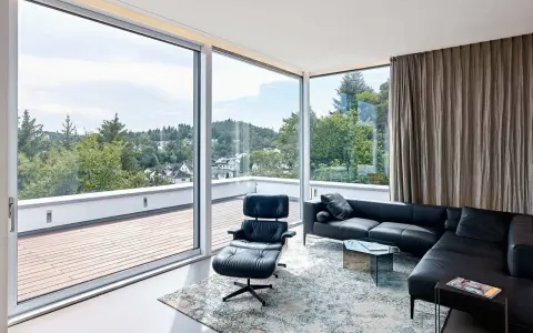 Nowoczesny salon z panoramicznym oknem narożnym, czarną skórzaną kanapą i fotelem Eames. Widok na zielone wzgórza.