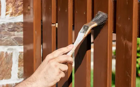 Ręka maluje drewniany płot pędzlem. Decyzja: podbitka lakierobejca czy impregnat?