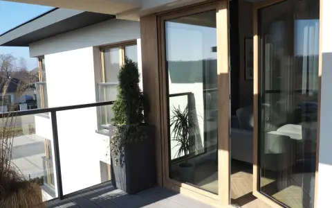 Nowoczesny balkon z drewnianymi, podwójnymi drzwiami balkonowymi. Okna dzielone poziomo wpuszczają dużo światła.
