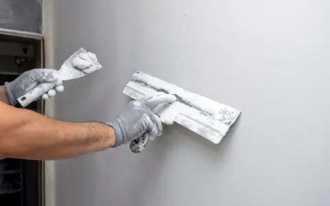 Ręce w rękawiczkach nakładają tynk na ścianę. Czas schnięcia tynku zależy od wielu czynników.