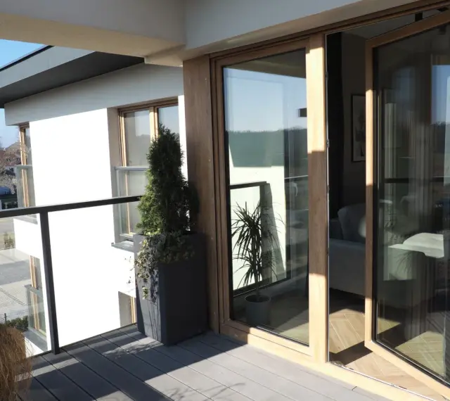 Nowoczesny balkon z drewnianymi, podwójnymi drzwiami balkonowymi. Okna dzielone poziomo wpuszczają dużo światła.