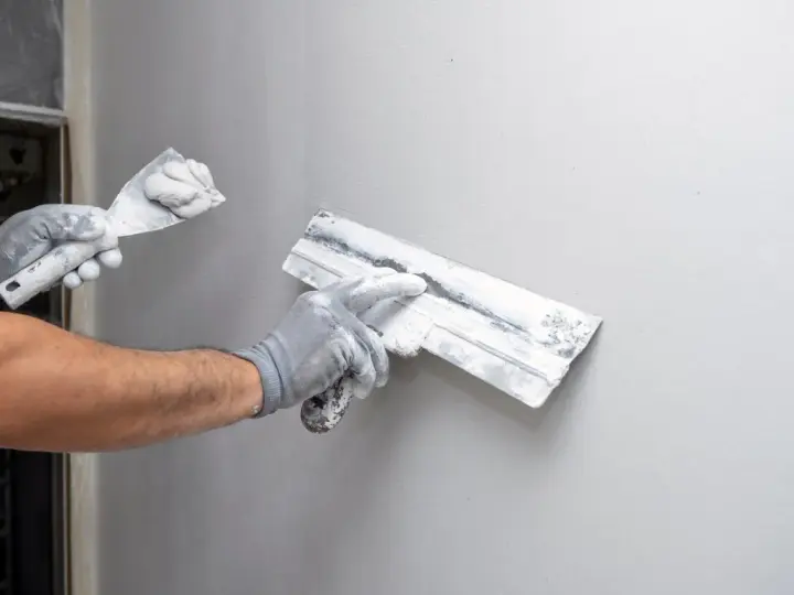 Ręce w rękawiczkach nakładają tynk na ścianę. Czas schnięcia tynku zależy od wielu czynników.
